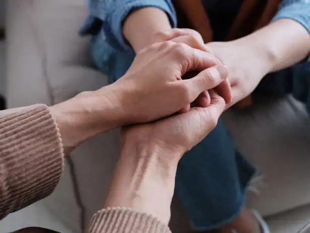Close nas mãos de duas pessoas sentadas uma de frente para a outra, em gesto de cuidado e apoio