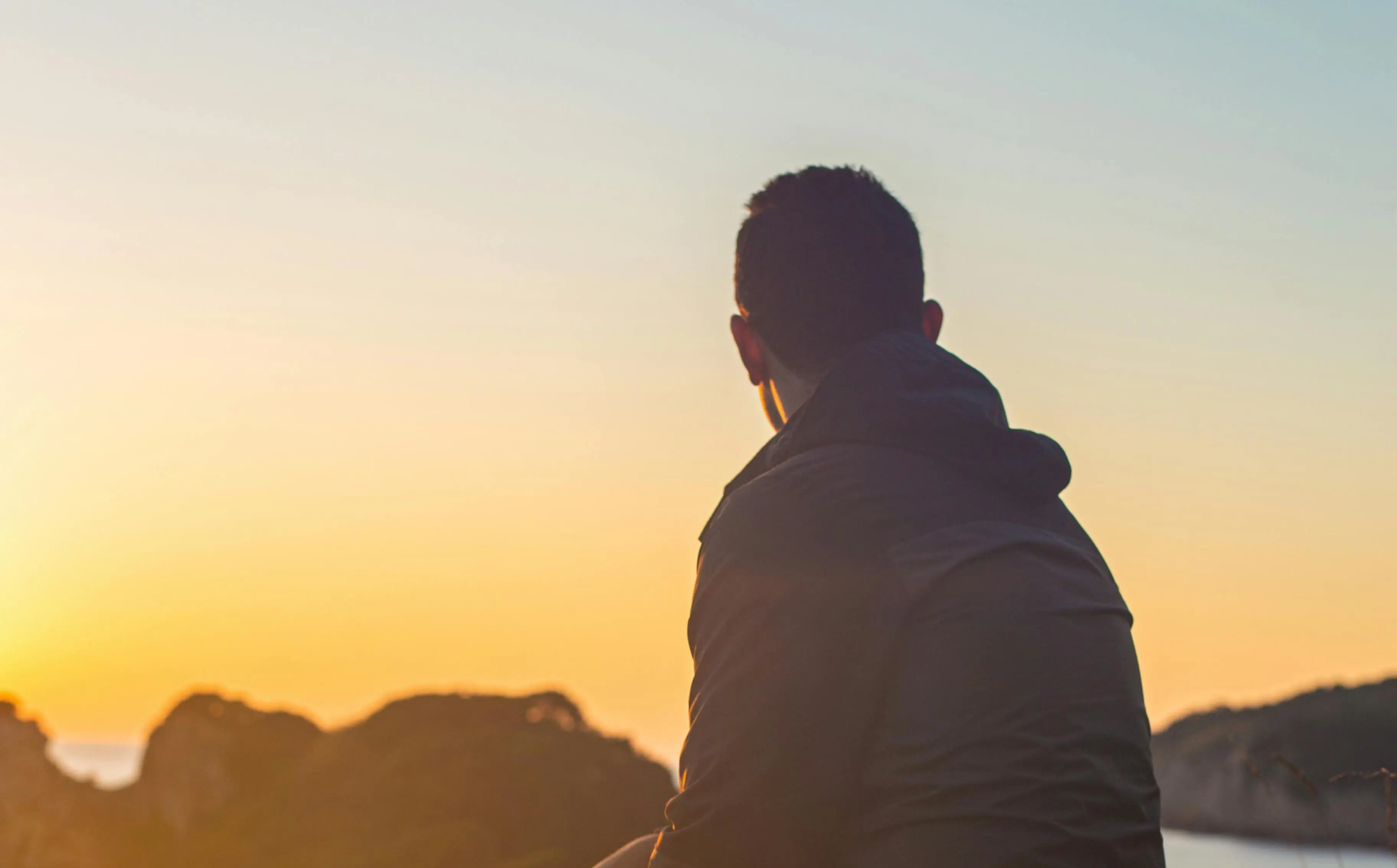 Homem sentado de costas olhando para o horizonte ao pôr do sol